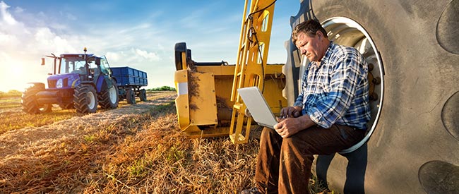 White middle aged farmer sitting in tire of large farm machine with his laptop in the afternoon of a blue sky day