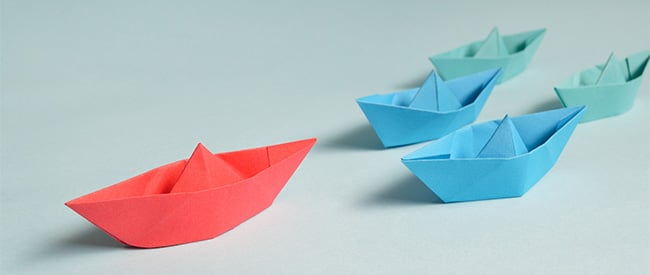 Origami boats, one red followed by two blue followed by two green on a pale grey background